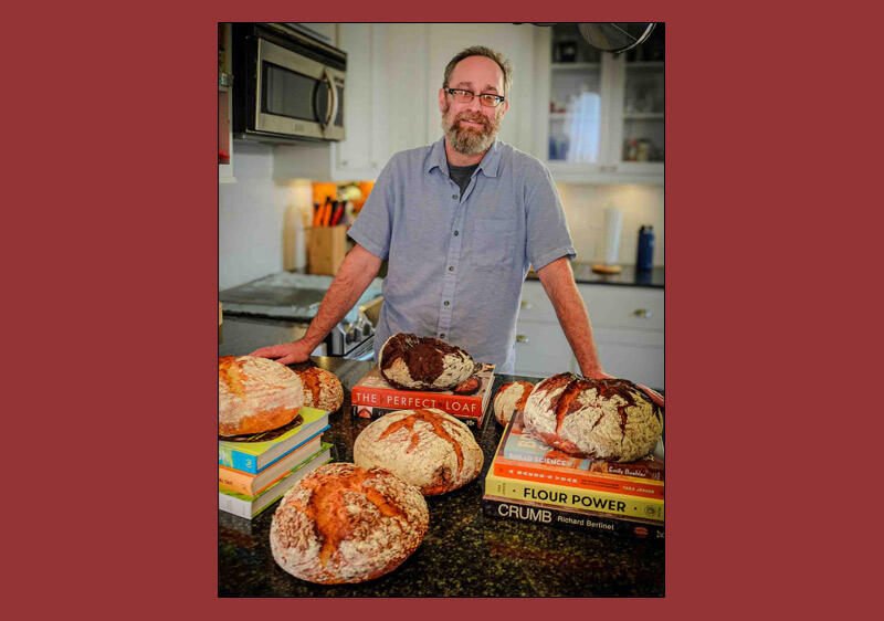 photo of a man with several fresh loaves of rustic looking breads and baking related books